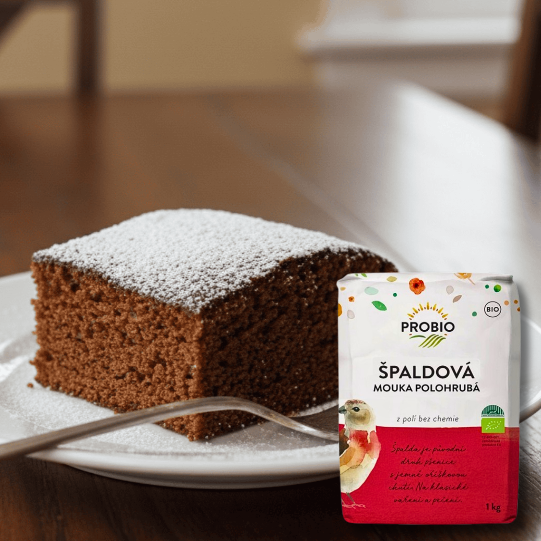 Slice of cocoa sponge cake dusted with powdered sugar on a white plate with a fork, next to a 1 kg bag of PROBIO organic spelt flour (semi-coarse).